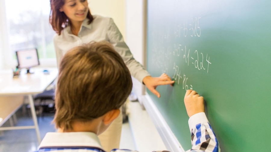 education, elementary school, learning, math and people concept - close up of little schoolboy with teacher writing on chalk board and solving task in classroom