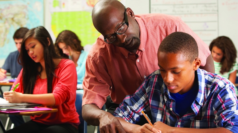 Teacher Helping Male Pupil Studying At Desk In Classroom